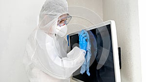 Portrait of woman afraid of viruses wearing protective medical suit and mask desinfecting TV with cleanser and detergent