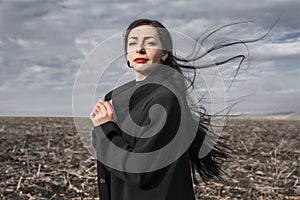 portrait of woman in a black coat whose hair is flying in the wind stands in a field