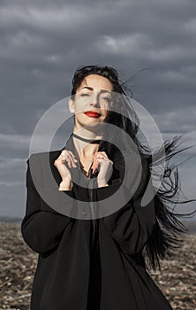 Portrait of woman in a black coat whose hair is flying in the wind stands in a field