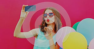 Portrait of woman blowing her red lips sending sweet air kiss taking selfie picture by smartphone with colorful balloons on pink