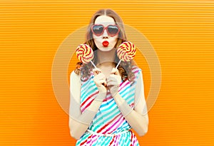 Portrait woman blowing red lips holding two lollipop in heart shaped sunglasses, colorful striped dress on orange wall