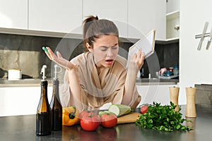 Portrait of woman cant cook, looking confused while making meal, holding recipe book, checking grocery list and staring