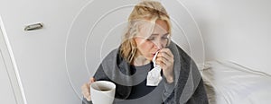 Portrait of woman catching a cold, staying home sick, drinking tea, sneezing in napkin, blowing nose, has covid symptoms