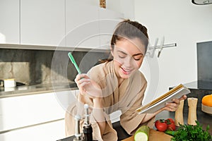 Portrait of woman checking grocery list, looking at vegetables, holding notebook, reading recipe while cooking meal in