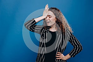 Portrait of woman with curly dark hair forgetting something, slapping forehead with palm and closing eyes  over blue