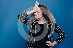 Portrait of woman with curly dark hair forgetting something, slapping forehead with palm and closing eyes  over blue