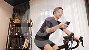 Portrait of woman cyclist with smartphone in hands training on indoor smart trainer. Female is cycling on stationary bicycle at ho