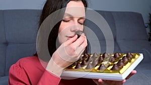 Portrait of woman is eating chocolate candies from box smelling them before it.