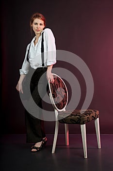 Portrait of woman, girl in man\'s suit with suspenders stands with chair.