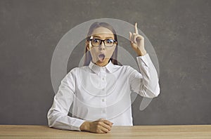 Woman in glasses sitting at a desk and pointing her finger up surprised by a smart idea