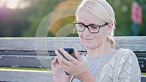 Portrait of a woman in glasses. Sits on the bench, looks at the smartphone screen