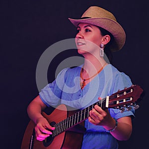 Portrait of a woman with a guitar in her hands on a black studio background. A female musician in a blue dress and a straw hat