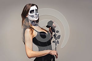 Portrait of woman with halloween skeleton makeup holding black rose flower over gray background