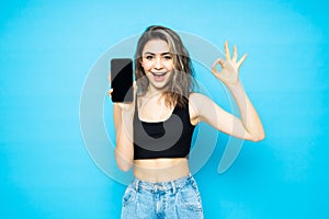 Portrait of woman holding blank screen mobile phone and showing ok gesture isolated over blue background