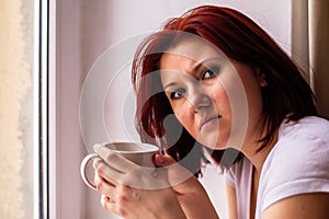 Portrait of woman holding white mug of coffee near window and looking at camera. Female has stressed and unhappy face. Copy space