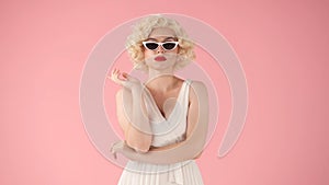 Portrait of a woman in the image of Marilyn Monroe wearing cat eye shape sunglasses in studio on pink background.