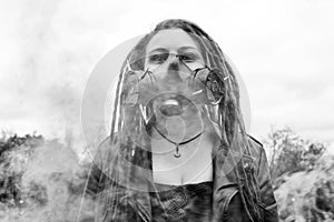 Portrait of a Woman in a jacket and dreadlocks in a gas mask with spikes. woman standing in smoke. Black and white photo