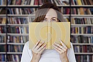 Portrait of woman in a library face half-covered by opened book, long hair. Young college student girl in co-working