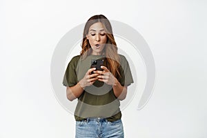 Portrait of woman looks surprised at mobile phone, say wow impressed, reading message, spot awesome deal online, store