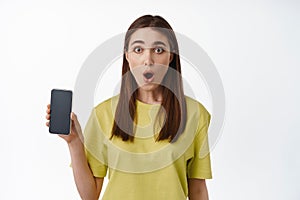 Portrait of woman looks surprised, shows interface, mobile phone screen app, demonstrate application on smartphone