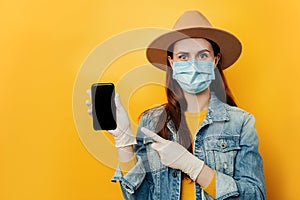 Portrait woman in medical face mask, points at smart phone, shows blank screen of gadget, wears hat, glowes and denim jacket