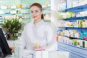 Portrait of woman pharmacist who is standing on her work place near cashbox in apothecary.