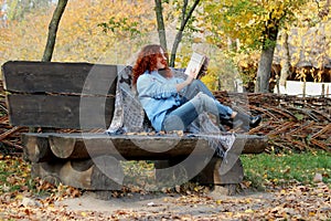 Beautiful Woman with red hair in the autumn park. sits on a bench and reads a book. Autumn background. Nearby is a warm blanket wi