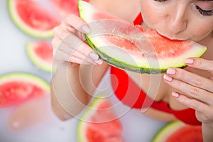 Portrait of a woman in a red swimsuit eating a watermelon and smirking. Redhead girl with red lipstick takes a bath with