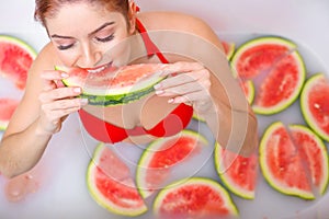 Portrait of a woman in a red swimsuit eating a watermelon and smirking. Redhead girl with red lipstick takes a bath with