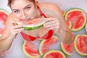 Portrait of a woman in a red swimsuit eating a watermelon and smirking. Redhead girl with red lipstick takes a bath with