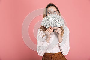 Portrait of woman 30s in trendy wear hiding while holding money fan isolated over pink background
