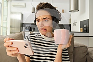 Portrait of woman with scared, concerned face watching video on smartphone, drinking tea, wearing headphones, sitting on