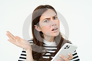 Portrait of woman shrugging, holding smartphone and looking confused at camera, isolated on white background