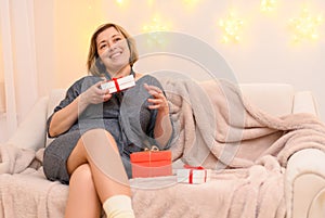 Portrait of a woman, sits on a couch and opens gifts, holiday lights on a wall