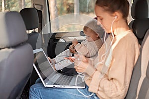 Portrait of woman sitting with notebook in car, her baby daughter in safety chair on backseat, female listening music on phone,