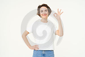 Portrait of woman smiling, showing okay sign with confidence, gives approval, recommends smth good, stands over white