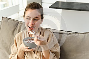 Portrait of woman on sofa in bathrobe, eating cereals with milk, watching tv and having breakfast
