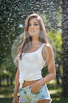 Portrait of woman in spray of water with white T shirt . She has a good gentle skin, sensual posture, and she smiles.