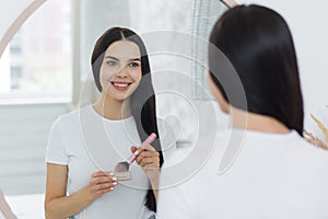 Portrait of a woman standing in front of a mirror and applying make-up with a brush on her face.