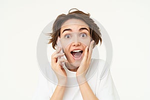 Portrait of woman with surprised face, answers phone call and looks excited, amazed by big news, stands over white