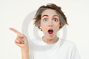 Portrait of woman with surprised face, showing promo to the left, pointing at something, standing over white background