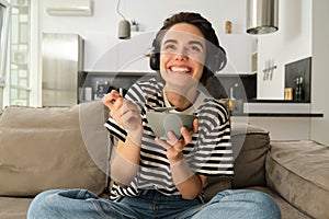 Portrait of woman watching tv in wireless headphones, looking at screen with interest and excitement, eating cereals in