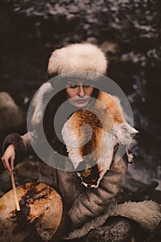 Portrait of a woman wearing fur hat and fox fur pelt on her shoulders.Playing shamanic drum in the forest.