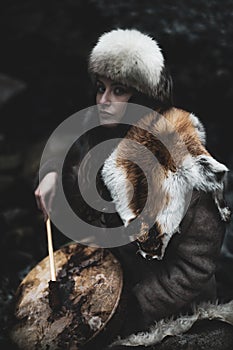 Portrait of a woman wearing fur hat and fox fur pelt on her shoulders.Playing shamanic drum in the forest.