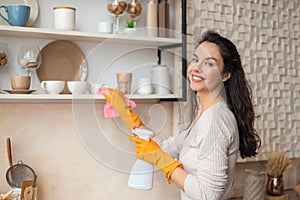 Portrait of woman wearing protective rubber gloves cleaning shelves in kitchen, holding spray and using microfiber cloth