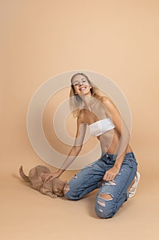 Portrait of woman wearing ripped jeans smiling petting peach colored cat with hand looking at camera on skin colored