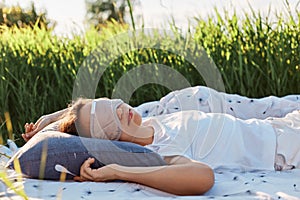 Portrait of woman wearing sleeping mask and white t shirt lying on soft bed in the middle of green field or meadow, female sleeps