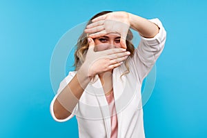 Portrait of woman in white jacket looking through photo frame made of hands. , blue background