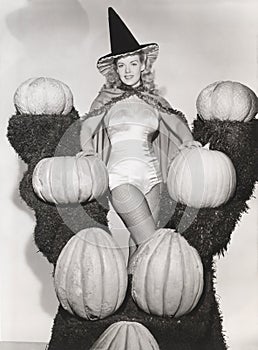 Portrait of woman in witch costume surrounded by pumpkins on grass shelves