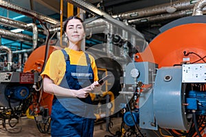 Portrait of a woman worker in a uniform and with a tablet in her hand. In the background is a boiler room. The concept